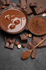 Wall Mural - glass bowl of chocolate cream or melted chocolate, pieces of chocolate and hazelnuts on dark concrete background