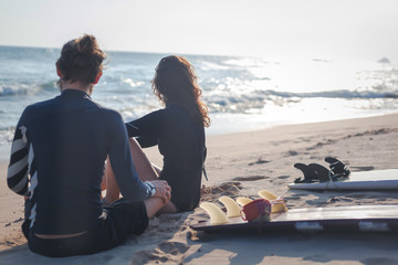 Wall Mural - Young beautiful loving couple friends are sitting on the sand by the ocean with surfboards and watching sports active lifestyle holidays honeymoon...