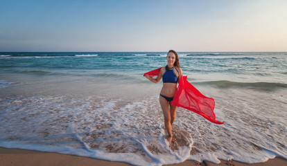 Wall Mural - Beautiful young brunette woman with long hair in a bikini with a bright red scarf in her hands on the ocean at sunset