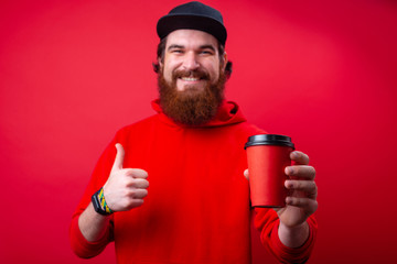 Bearded guy  in rd hoody with toothy smile holding a cup and showing thumb up over isolated red background