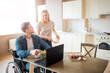 © estradaanton - Young worker sit on wheelchair and look at woman. He work with laptop and eat salad. They look at each other and smile. Woman stand beside guy. Happy couple together.