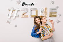 Portrait Two Attractive Joyful Models With Stylish Makeups, Luxury Coiffures Having Fun, Drinking Champagne On White Background With Inscription Beauty Zone . Expressing True Positive Emotions, Beauty