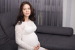 © sementsova321 - Indoor shot of young beautiful pregnant woman with dark wavy hair, sitting on comfortable gray sofa at window in modern apartmet, wearing white dress, looks at camera. Motherhood and pregnancy concept