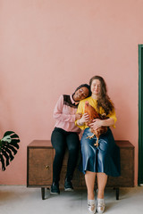 Young interracial female couple sits together with beloved domestic friend - cock and hugs each other on pink wall background. Lifestyle portrait of unusual female pet lovers. Odd f female friendship.