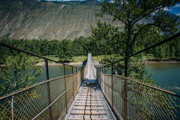 Suspension bridge over the mountain river. The hanging bridge surrounded by lush green trees on the background of mountains and white clouds on the blue sky. Travelling to Russia.