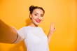 Portrait of charming girlish funky hipster student posing making selfie v-sign sharing photo posting on her social network account wearing white cotton clothing isolated over vivid background