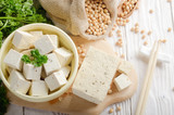 Fototapeta  - Soy Bean curd tofu in clay bowl and in hemp sack on white wooden kitchen table. Non-dairy alternative substitute for cheese. Place for text