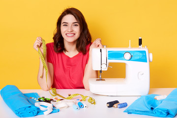 Close up portrait of charming woman seamstress sitting at table with sewing machine on yellow background in studio, dressmaker sews new dress, looks happy, holds measure tape, designer makes outfit.