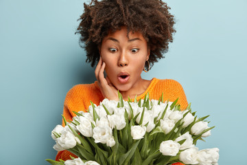 Astonished Afro American lady stares at white beautiful flowers, can not believe eyes, keeps hand on cheek, wears orange sweater, isolated over blue background. People and unexpected reaction concept