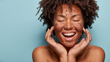 Cheerful Dark Skinned Lady Applies Coffee Scrub On Face, Pampers Skin, Closes Eyes From Pleasure, Smiles Positively Has Bare Shoulders, Stands Against Blue Background, Free Space Aside. Beauty Concept