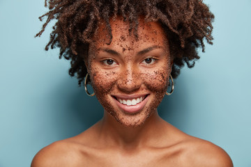 Wall Mural - Beautiful mixed race female has skin scrub on face, smiles gently and looks directly at camera, makes cosmetic masks from coffee, has curly hairstyle, bare shoulders, isolated over blue background