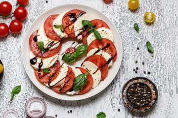 Wall Mural - Italian caprese salad with ripe tomatoes, fresh basil and mozzarella cheese on grey stone background. Italian caprese salad in white plate with sliced tomatoes