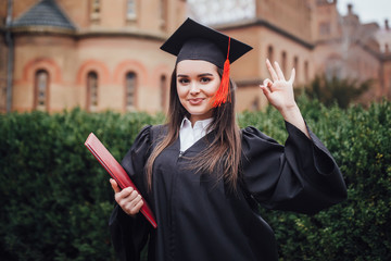 Wall Mural - Portrait of female college student in graduation cap and gown on campus with okay sign!