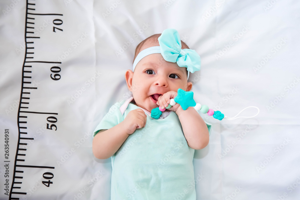 A three month baby in mint green clothes lying on a bed on which a measuring ruler for growth is drawn. Teething tool in hands, chewing - obrazy, fototapety, plakaty 