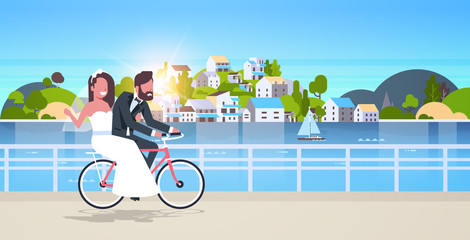 Wall Mural - just married man woman riding bicycle romantic couple bride and groom cycling bike having fun wedding day concept mountain city island sunset background full length horizontal flat