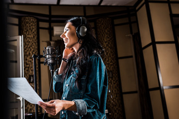 Wall Mural - selective focus of pretty woman holding paper with text while singing in recording studio