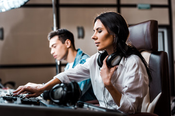 Wall Mural - selective focus of pretty sound producer in earphones working at mixing console near mixed race friend