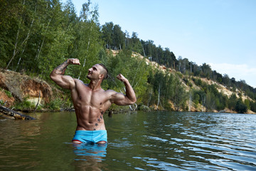 Gorgeous guy stands in the pose of bodybuilder in the lake on the background of a beautiful sandy ravine and green forest and looks smiling to the side