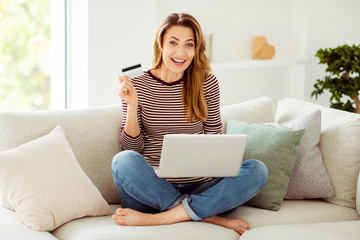 Portrait of her she nice-looking attractive lovely cheerful cheery glad wavy-haired girl sitting on divan lotus position showing new card sale discount in light white interior room