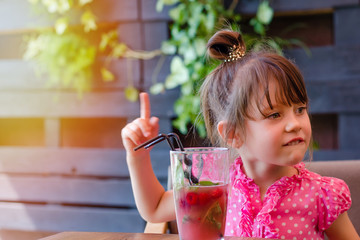 Adorable little girl drinking lemonade with raspberry and basil at table in cosy outdoor cafe. Happy and healthy childhood concept. Copy space