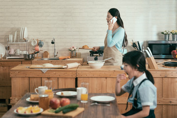 Wall Mural - ignore family gadget addiction. busy asian wife mother holding wooden plate with croissant walking in modern kitchen talking with husband on cellphone in morning. little girl eating having breakfast