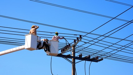 Two electrians on electric cable car lifts are working to install electrical transmission on power pole with many cable lines against blue clear sky background in sunny day