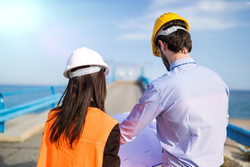 Ingegnere uomo e donna di spalle stanno guardando il progetto sul foglio a2 di carta prima di valutare i lavori per la costruzione del ponte sl mare. Cancello d'ingresso al cantiere sullo sfondo.