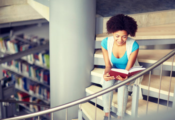 education, high school, university, learning and people concept - smiling african american student girl reading book sitting on stairs at library