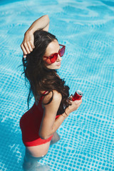 Wall Mural - Fashionable and happy brunette model woman with a perfect sexy figure in stylish red swimsuit and trendy red sunglasses in the swimming pool holding a cold drink in her hand