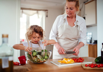 Wall Mural - A portrait of small girl with grandmother at home, preparing vegetable salad.