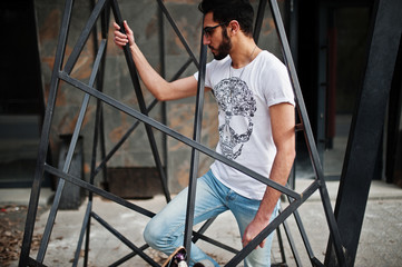 Wall Mural - Street style arab man in eyeglasses with longboard posed inside metal pyramid construction.