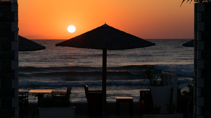 sunrise on the seafront with reed sun silhouette umbrellas, the sea with waves and dinner table.