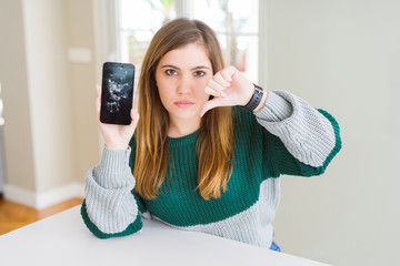 Canvas Print - Beautiful young woman holding broken smartphone with angry face, negative sign showing dislike with thumbs down, rejection concept