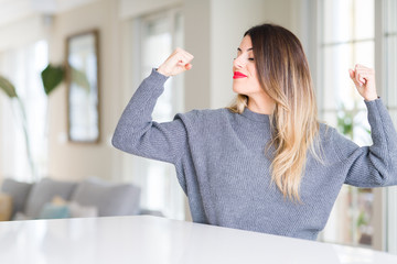 Young beautiful woman wearing winter sweater at home showing arms muscles smiling proud. Fitness concept.