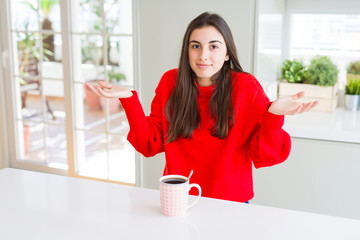 Canvas Print - Beautiful young woman drinking a cup of black coffee clueless and confused expression with arms and hands raised. Doubt concept.