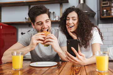 Canvas Print - Picture of european using mobile phone while eating hamburger during breakfast in kitchen at home