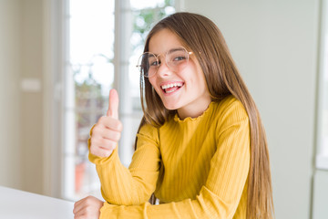 Beautiful young girl kid wearing glasses doing happy thumbs up gesture with hand. Approving expression looking at the camera showing success.