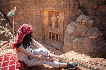 Asian woman traveler sitting on carpet viewpoint in Petra ancient city looking at the Treasury or Al-khazneh, famous travel destination of Jordan and one of seven wonders. UNESCO World Heritage site.