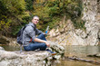 © Rithor - Side view - a young guy tourist in daily clothes with a backpack drinks water from a clean river in the mountains. Concept of active recreation and rehabilitation