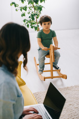 Wall Mural - kid sitting on rocking horse while mother working with laptop in living room