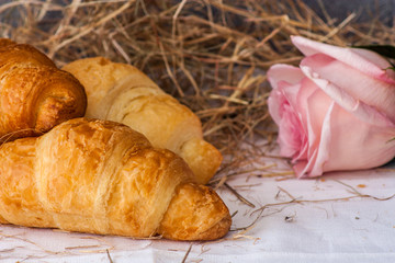 Three crisp fresh croissants and a pink rose flower close-up on a white towel against a background of hay and a gray wall.