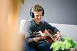 © Westend61 - Smiling young man with headphones sitting on couch playing guitar