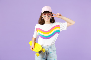 Wall Mural - Pretty teen girl in vivid clothes heart eyeglasses hold yellow skateboard showing victory sign isolated on violet pastel wall background. People sincere emotions lifestyle concept. Mock up copy space.