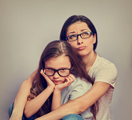 Wall Mural - Sad young casual mother looking up and hugging her cute unhappy offended thinking kid girl on violet wall background. Family in fashion eye glasses.