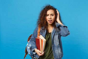 Young sad african american girl teen student in denim clothes, backpack hold books isolated on blue background studio portrait. Education in high school university college concept. Mock up copy space.