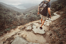 Man With Backpack And Camera Is Hiking By The Stone Path In The Beautiful Nature. There Are Fog And Mountains At Background.