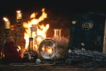 Crystal ball, magic book, magic potion and other wizard accessories on a table on a burning fire background.