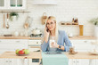© oes - Freelance concept. Beautiful business woman in casual clothes and glasses is examining documents and smiling while working with a laptop in kitchen. Working at home