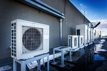 Square air-conditioning unit on the roof with a round fan. In the background gradually receding other units that are out of focus. On the right side light blue sky and commercial space.