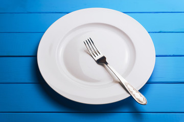 Poster - White plate and a fork with a spoon on a blue wooden table background.